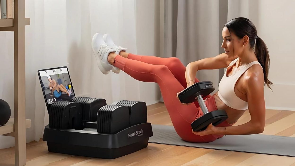 A fit woman in a white sports bra and coral leggings performs a Russian twist exercise using an adjustable dumbbell in a modern home gym. She is seated on a yoga mat, balancing on her tailbone with legs elevated. A NordicTrack adjustable dumbbell set is placed beside her, and a tablet screen in the background displays a virtual workout session. The setting is bright and stylish, with neutral-colored curtains and wooden flooring.