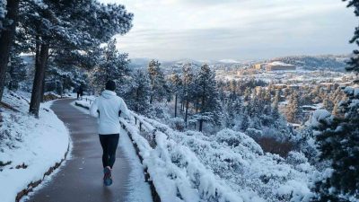 A runner dressed in cold weather running gear, including a white thermal jacket, black running tights, gloves, and a beanie, jogging along a snow-covered scenic trail. The winding path is lined with snow-dusted trees, overlooking a winter landscape with hills and a city skyline in the distance. The crisp winter atmosphere and runner's breath in the cold air highlight endurance and determination in cold weather conditions.