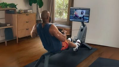 A muscular man wearing a sleeveless blue shirt and orange shorts is using a modern rowing machine in a well-lit home gym. He is engaged in an interactive rowing workout displayed on a large digital screen. The room features wooden flooring, a dresser, and indoor plants, creating a cozy workout environment.