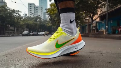 A close-up of a runner’s foot wearing a bright yellow and orange Nike running shoe with a green swoosh, paired with white Nike socks, stepping on an urban street. The background features blurred city buildings, trees, and passing cars, emphasizing the shoe’s design and motion.
