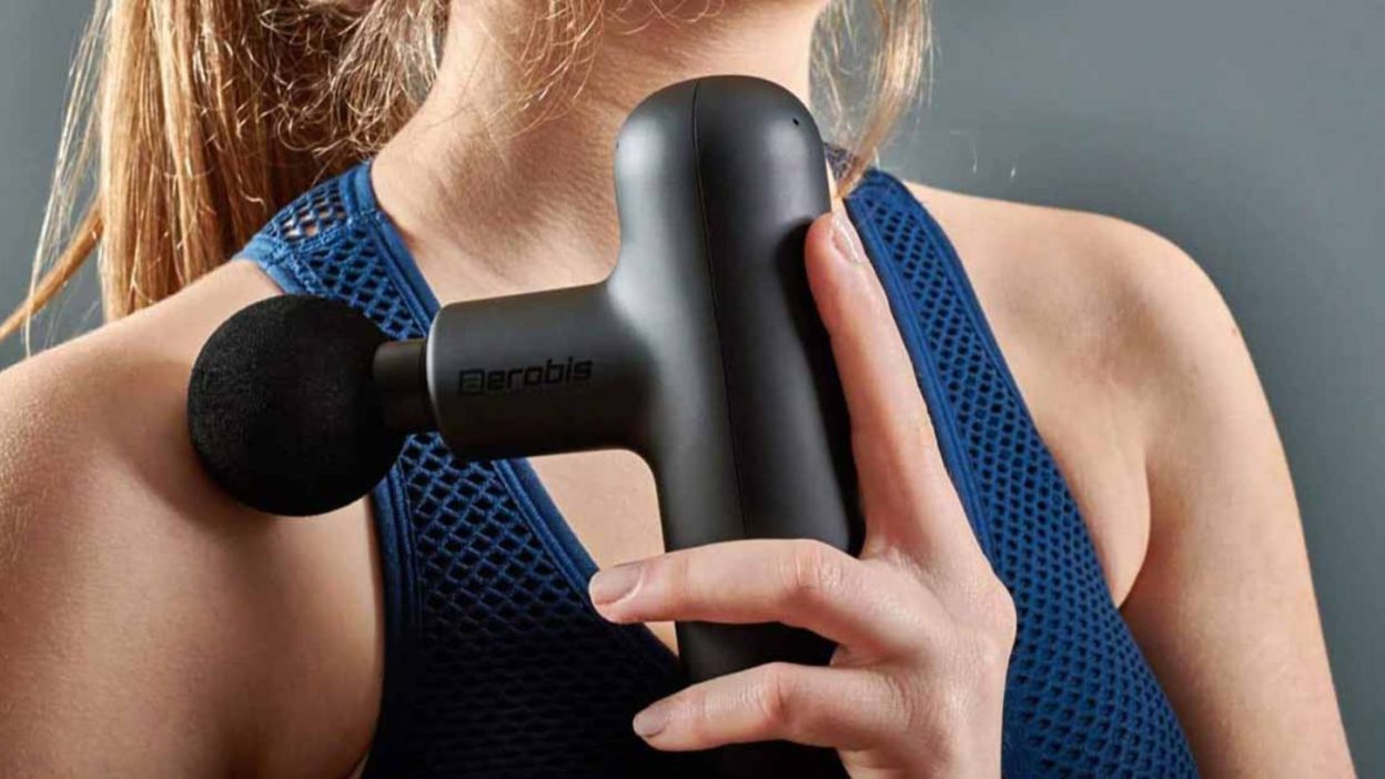 A close-up of a woman using a black Aerobis massage gun on her shoulder. She is wearing a blue athletic tank top, and the massage gun is positioned against her upper arm. The background is a neutral gray, emphasizing the focus on the massage device and its use for muscle recovery.