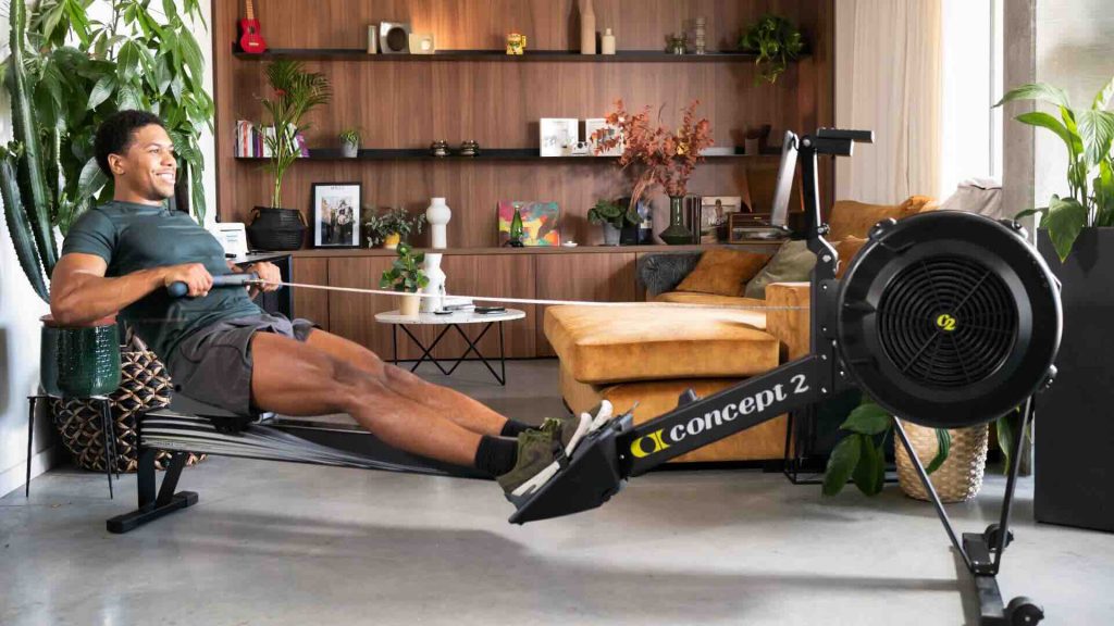 A fit man in a dark green athletic shirt and gray shorts is using a Concept2 rowing machine in a stylish living room with modern wooden shelves, indoor plants, and a cozy mustard-colored sofa. He is smiling while rowing, demonstrating a home workout routine.