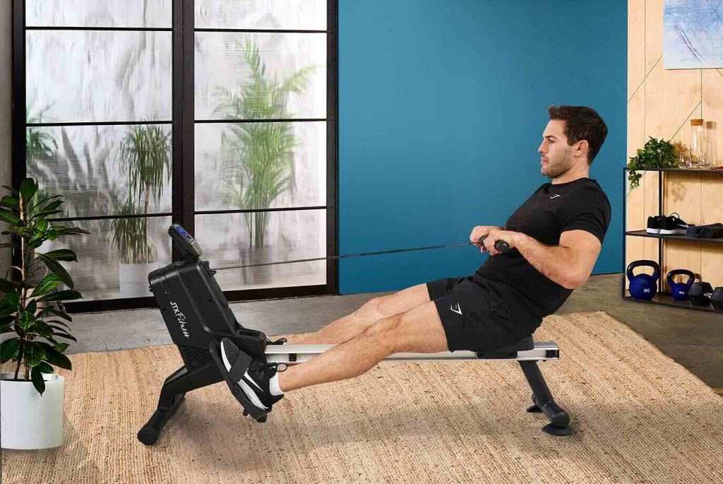 A fit man in black athletic wear is using a magnetic rowing machine in a modern home gym with a blue accent wall. He is seated on a beige rug, pulling the rowing handle with controlled form. The gym space features kettlebells, plants, and minimalist decor, creating a clean and motivating workout environment.